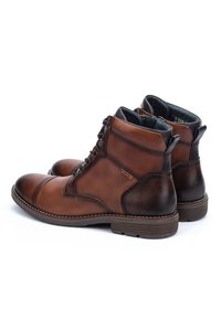Botas de cuero marrón hasta el tobillo con cordones en la parte delantera, paneles laterales texturizados, acentos de tacón marrón oscuro y tiradores, sobre fondo blanco.