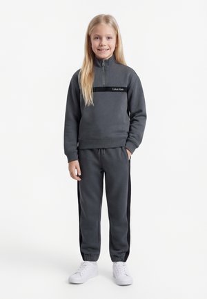 Κορίτσι που στέκεται και χαμογελά, φορώντας μια σκούρα γκρι ζακέτα με φερμουάρ Calvin Klein και ταιριαστό παντελόνι φορμάς με λευκά αθλητικά παπούτσια.