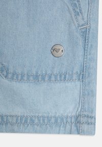Lyseblå denimstoff med synlige sømmer og et sirkulært metallaccenter med et diamantmønster på framsiden av lommen.