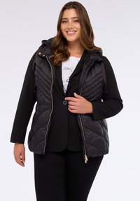 Černá prešívaná vesta s kapucí, s diagonálním prošíváním, zlatými doplňky a předním zipem. Nositá přes černý blazer a bílý tričko.