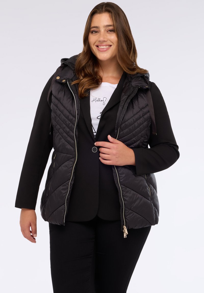 Černá prešívaná vesta s kapucí, s diagonálním prošíváním, zlatými doplňky a předním zipem. Nositá přes černý blazer a bílý tričko.