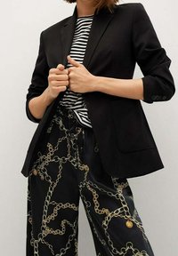 Blazer noir au design ajusté, fermeture à un bouton et revers crantés, associé à un pantalon noir avec un motif de chaîne dorée.
