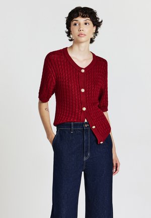Junge Frau mit kurzen, lockigen Haaren, die eine rote, geknöpfte Strickjacke und dunkelblaue, hochgeschnittene Schlagjeans trägt, steht mit den Händen in den Taschen.
