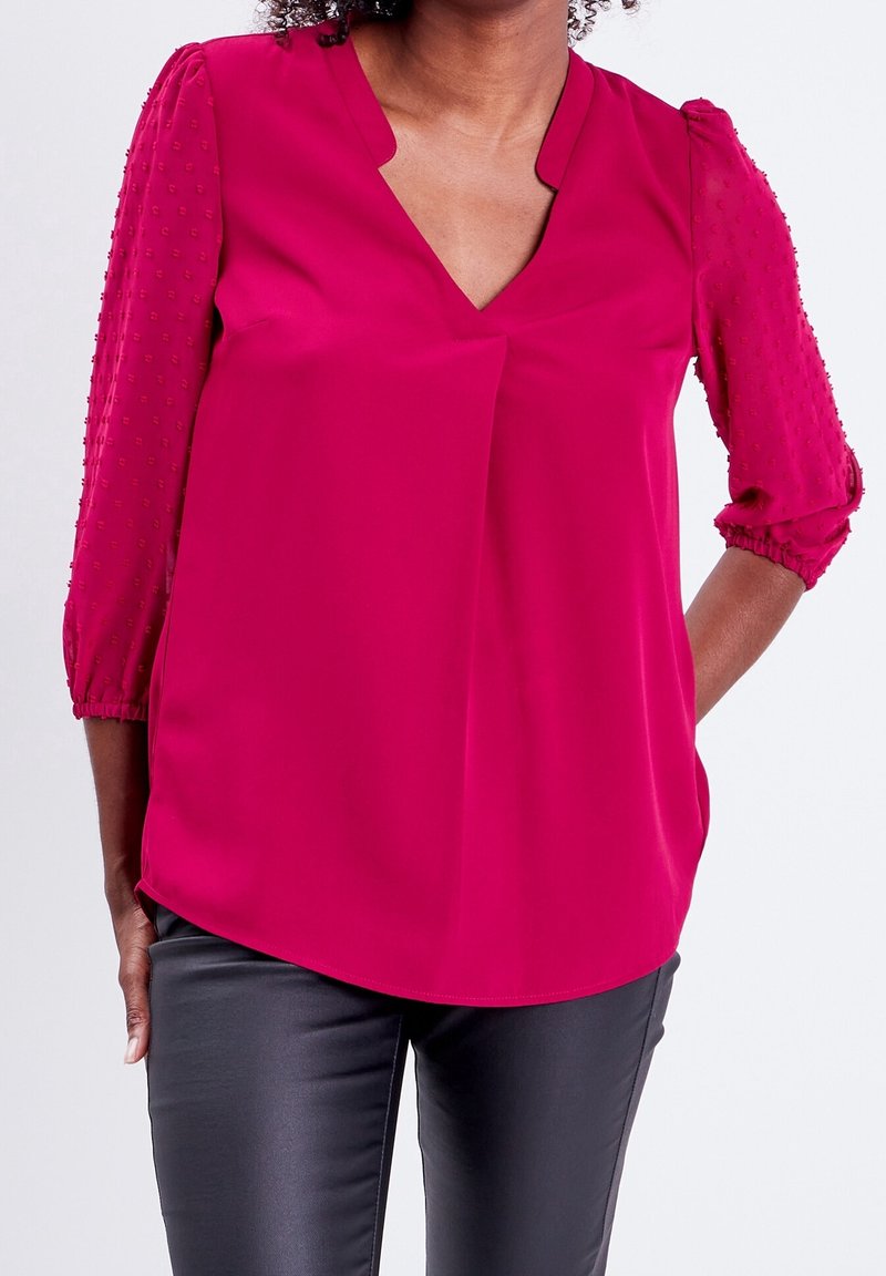 Blusa fucsia con escote en V y mangas 3/4 abullonadas con puntos en relieve, combinada con pantalones negros ajustados.