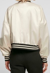 Veste bomber courte en satin crème avec ourlet froncé, ornée de poignets et d'une taille côtelés rayés noirs et beiges, texture lisse.