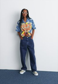 Blusa gráfica de manga corta en azul con estampado multicolor, anudada a la cintura, combinada con jeans de pierna ancha en azul oscuro y zapatillas beige.