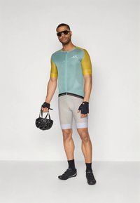 Homem em jersey de ciclismo azul-água e amarelo, calções cinzentos de ciclismo, luvas pretas e sapatos, segurando um capacete preto, usando óculos de sol grandes, de pé sobre um fundo branco.