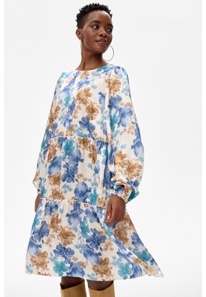 Kobieta ubrana w sukienkę z długim rękawem w kwiatowy wzór w kolorach niebieskim, brązowym i zielonym, do tego pasuje jasnobrązowe botki i duże kolczyki koła.