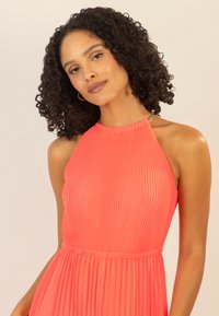 Un vestido halter de plisados en un brillante color coral, con una cinturilla fruncida y un sutil acento en hardware dorado en el escote. Textura suave en toda la prenda.