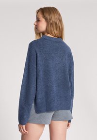 Niebieski sweter dzianinowy w luźnym kroju, z prążkowanymi mankietami i dolnym brzegiem oraz okrągłym dekoltem, w zestawie z jasnoszarymi szortami.