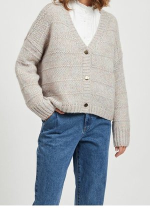 Frau trägt eine beige grob gestrickte Strickjacke mit großen Knöpfen über einer weißen Bluse, kombiniert mit hoch taillierten blauen Jeans.