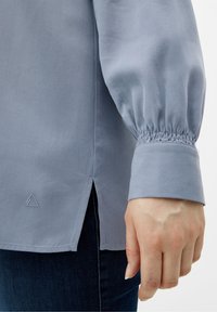Himmelbluse aus weichem Stoff mit gepufften Ärmeln und elastischen Bündchen. Verfügt über seitliche Schlitze und ein kleines dreieckiges Logo am Saum.
