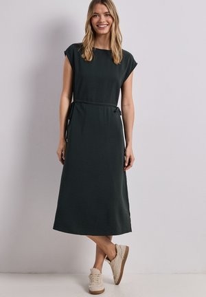 Femme souriante aux longs cheveux blonds, portant une robe midi sans manches vert foncé et des baskets beiges, debout contre un mur uni.