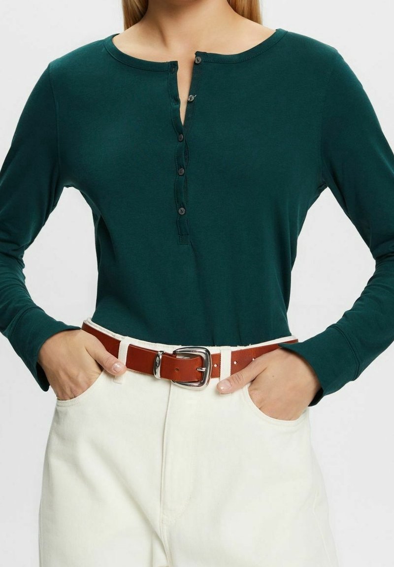 Langärmliges grünes Henley-Shirt mit rundem Halsausschnitt, vorderen Knöpfen, kombiniert mit hochtaillierten weißen Hosen und einem braunen Ledergürtel.