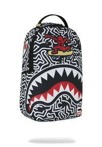 Mochila negra con patrón de laberinto blanco, diseño de boca de tiburón roja y una figura amarilla de skate. Cuenta con un asa en la parte superior y cierre de cremallera.