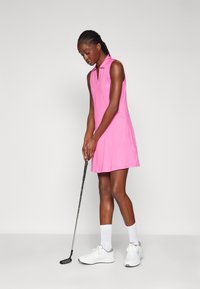 Abito da golf senza maniche di colore rosa con colletto, orlo a ginocchio e tasche laterali. Indossato con sneakers bianche e calze alte fino al ginocchio, mentre tiene un bastone da golf.