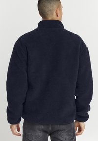 Sötét tengerészkék fleece pulóver. Puha, texturált anyag. Magas gallér és laza fit. Figyelemre méltó varrások a varratok mentén.