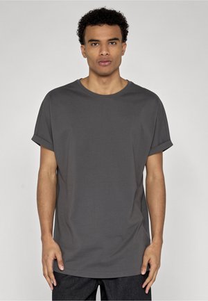 Homme portant un t-shirt uni gris foncé à manches courtes avec les manches retroussées, debout devant un fond clair uni.