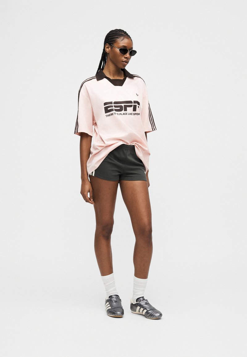 Femme portant un maillot de sport ESPN rose oversize, un short noir, des chaussettes blanches, des baskets argentées et des lunettes de soleil, debout devant un fond uni.