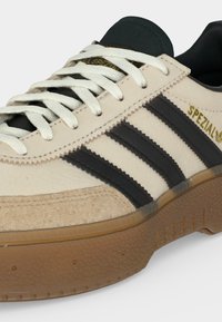 Κρεμ και μαύρο αθλητικό παπούτσι Adidas Spezial με λευκά κορδόνια, καφέ υφασμένο πέλμα και τρεις μαύρες ρίγες στο πλάι.