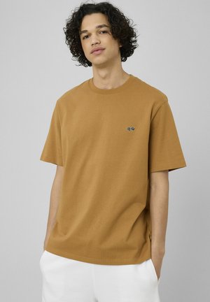 Jeune homme aux cheveux bouclés portant un t-shirt marron à manches courtes et un pantalon blanc, debout les mains dans les poches devant un fond uni.