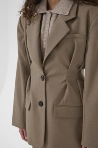 Ανοιχτό καφέ σακάκι με ραμμένες λεπτομέρειες και δομημένους ώμους, δύο κουμπιά, εμπρόσθιες τσέπες και λείο ύφασμα. Φοριέται πάνω από ένα μπεζ τοπ με γιακά.