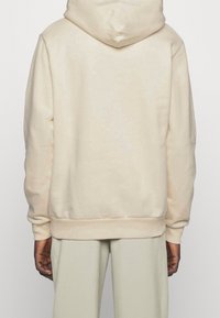 Sudadera con capucha de color crema con bolsillo canguro, puños y dobladillo acanalados. Cuenta con capucha con cordón y una textura suave y lisa.