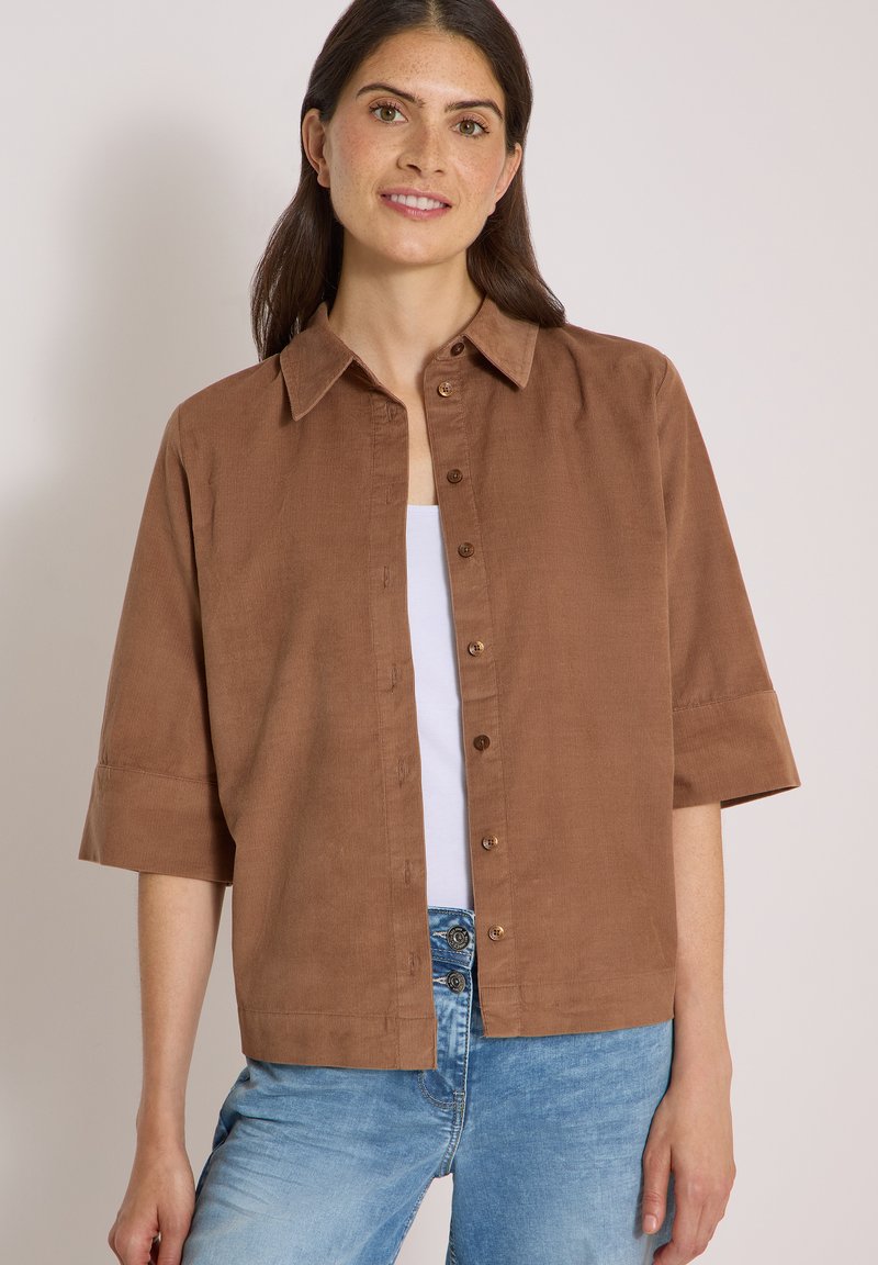 Femme aux longs cheveux foncés portant une chemise marron ample à boutons sur un haut blanc et un jean bleu clair, debout devant un fond neutre.