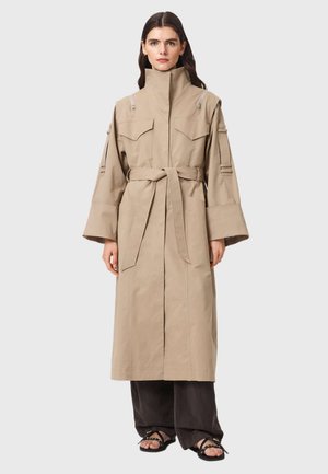 Vrouw draagt een beige lange trenchcoat met brede mouwen en ceintuur over donkere wijde broekspijpen en zwarte sandalen, staand tegen een effen achtergrond.