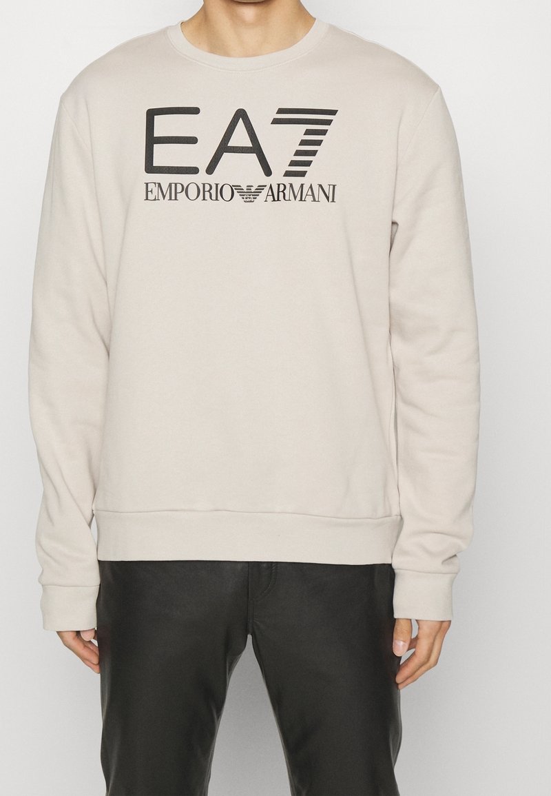 Světlá béžová mikina z měkké látky, s kulatým výstřihem, dlouhými rukávy a černým potiskem loga přes hrudník.