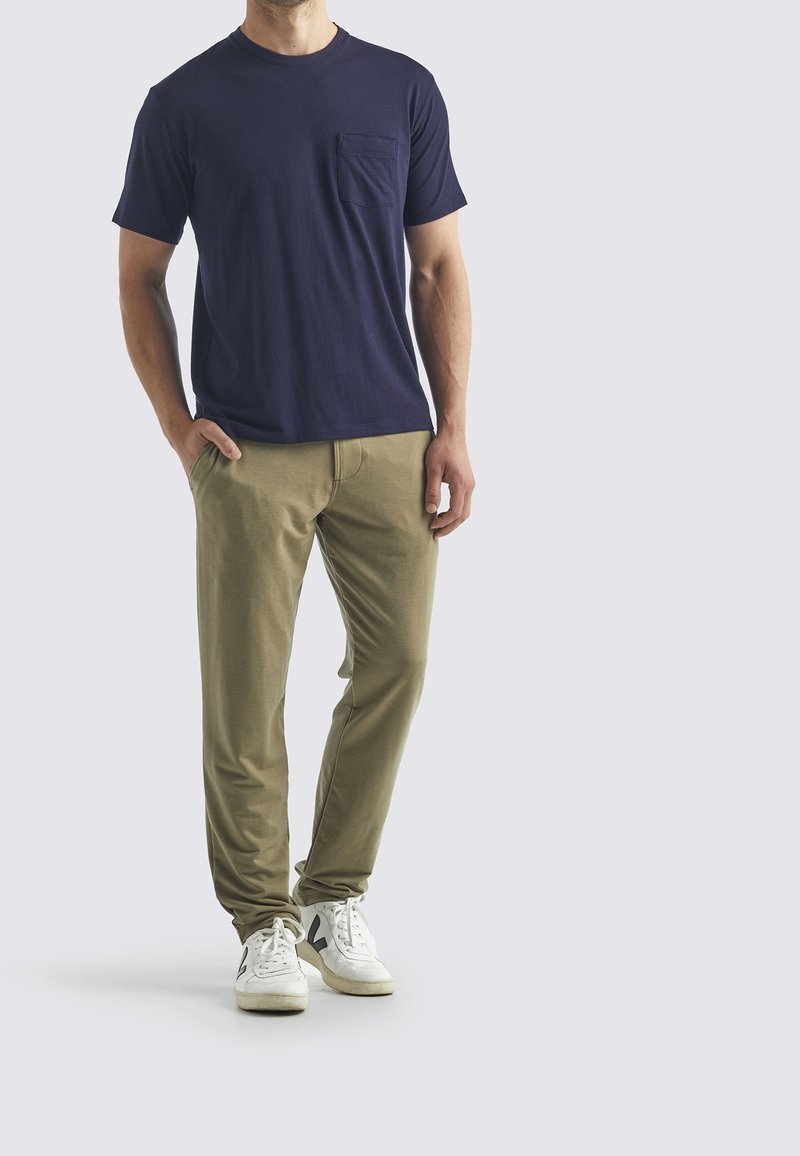 Man som bär en marinblå kortärmad skjorta med bröstficka, olivgröna byxor och vita sneakers, står med en hand i fickan.