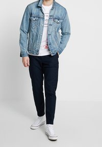 Chaqueta de mezclilla con un lavado azul medio, botones en la parte delantera, dos bolsillos en el pecho, camiseta blanca debajo, pantalones oscuros y zapatillas blancas.