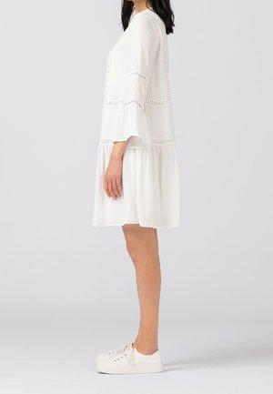 Vestido blanco con mangas de tres cuartos acampanadas y detalles de ojales, con fondo acampanado, combinado con zapatillas blancas, visto de lado.