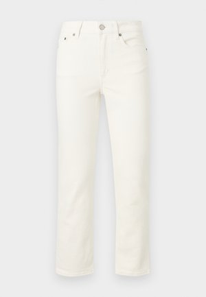 Γυναίκα φορώντας μαύρη μπλούζα μακρυμάνικη με γιακά, λευκό cropped παντελόνι και μαύρα μυτερά παπούτσια με αγκράφα, στέκεται σε απλό φόντο.