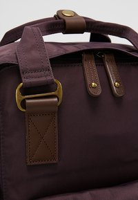 Sac fourre-tout en tissu violet avec des accents en cuir marron. Il comporte une poignée supérieure, des éléments en métal doré, et deux points d'attache en cuir cousus.