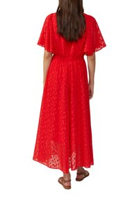 Vrouw in een rood gemusterde midi-jurk met vlindermouwen en smocktail, gecombineerd met bruine platte sandalen, staande met haar rug naar de camera gekeerd.