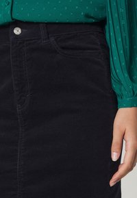 Gros plan d'une personne portant un pantalon en velours côtelé noir et un chemisier vert texturé à manches longues, avec une main posée à côté du pantalon.