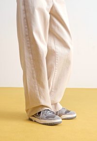 Beige vide bukser laget av et teksturert stoff, kombinert med grå joggesko med svarte detaljer og hvite såler, på et gult gulv.