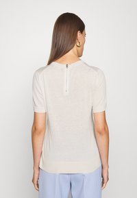Světle béžový top s krátkým rukávem a kulatým výstřihem, který má zip na zadní straně a hladkou jemně pletenou texturu.