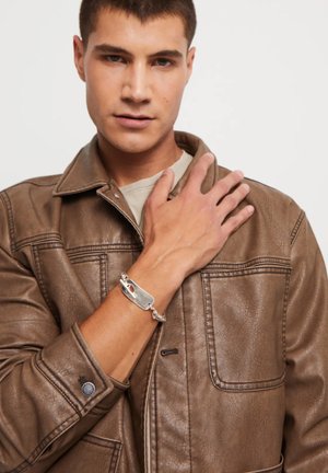 Chaqueta de cuero marrón con cuello sobredimensionado, bolsillos frontales y una pulsera plateada con un diseño de cierre único, llevada en una mano que descansa sobre la chaqueta.