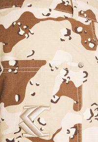 Kamouflage-mönstrad tyg i beige och bruna toner med en texturerad yta. Har en synlig metallknapp och en broderad logotyp.