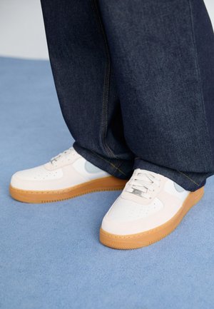 Baskets blanches à semelles en gomme portées avec un jean large foncé sur un sol recouvert d’un tapis bleu clair.