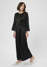 Frau in einem langen schwarzen Satin-Kleid mit langen Ärmeln und einer verdrehten Detail an der Taille, kombiniert mit beigen Slingback-Schuhen mit niedrigem Absatz.