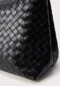 Bolsa tecida de couro preto com um padrão texturizado e acolchoado, apresentando uma forma suave e flexível. Superfície lisa e brilho sutil em evidência.