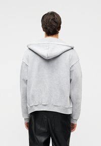 Άνδρας που στέκεται πλάτη, φοράει ανοιχτό γκρι φούτερ με κουκούλα και μαύρο παντελόνι με υφή, σε απλό λευκό φόντο.