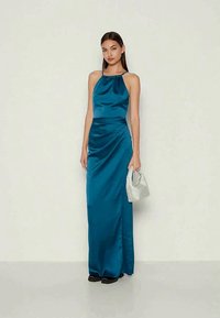 Vestido de noite em cetim azul teal com decote halter e detalhes franzidos na lateral. A modelo segura uma clutch de lantejoulas prateadas, vestindo sapatos pretos.