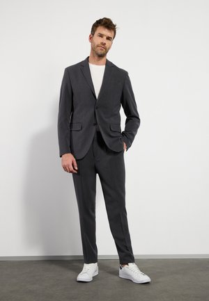 Giacca e pantaloni di un elegante grigio scuro realizzati in un tessuto liscio, abbinati a una t-shirt bianca e sneakers bianche. Linee pulite e design slim.