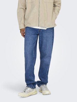 Personne portant une veste beige clair, un jean bleu droit et des baskets de randonnée beiges et grises, debout devant un fond uni.