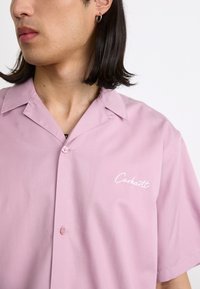 Lichtroze korte mouwen shirt met een knoopsluiting aan de voorkant, met een geborduurd wit "Carhartt" logo en een puntkraag.