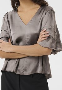 Sametová, taupe halenka s výstřihem do V, sklady na rukávech a volným střihem. Design zahrnuje vrstvený vzhled a hladkou texturu.